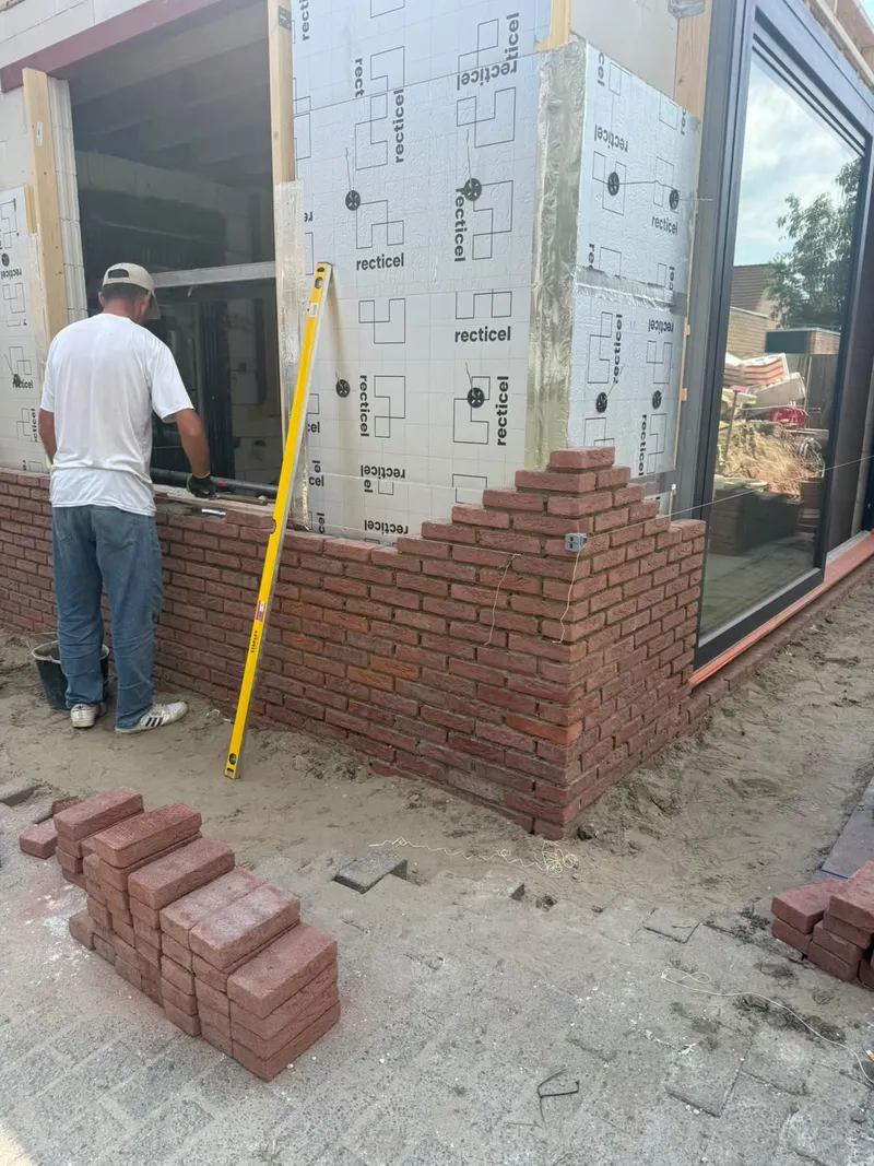 Vakman stelt een kozijn af met een waterpas terwijl het metselwerk van de aanbouw wordt opgetrokken