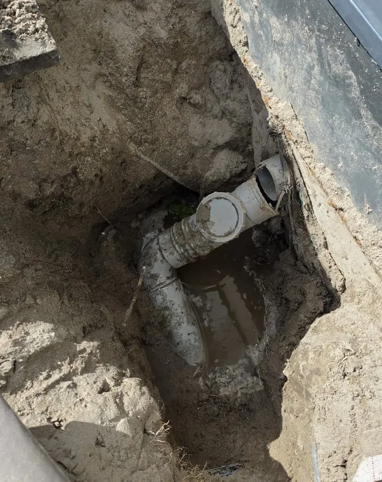 Opgegraven oude grijze afvoerleiding met bocht en T-stuk naast een donkere gevel, in een kuil met water