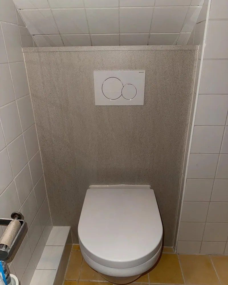 Hangend toilet met bedieningsplaat tegen een wandpaneel in natuursteenlook, onder een schuin plafond