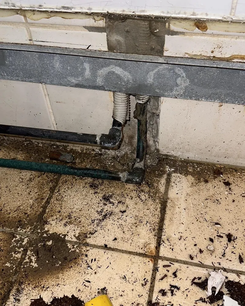 Gestripte badkamerwand met stalen frame en oude leidingen boven een vervuilde tegelvloer
