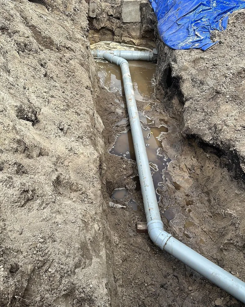 Lichtgrijze PVC-rioolleiding met bochten in een uitgegraven sleuf met grondwater