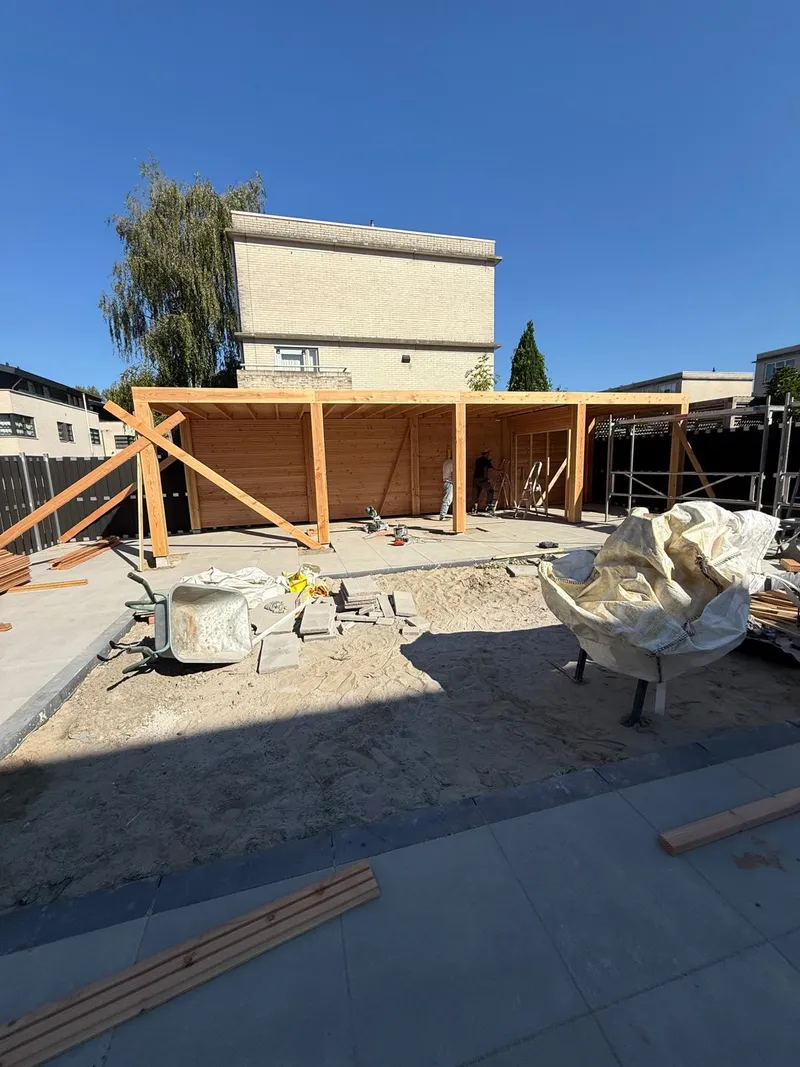 Houten overkapping in aanbouw op een nieuw terras, met staanders, dakbalken en een houten achterwand
